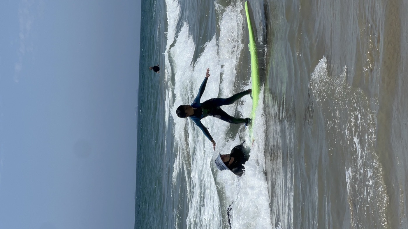 A young student standing up on a soft-top surfboard for the first time, coach beside them in the whitewater